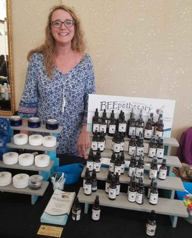 Laurie smiling in a blue floral dress standing behind BEEpothecary product display.