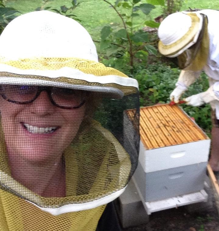Laurie smiling with a beekeepers hat and net on with Pete in the background tending to honey bee box.