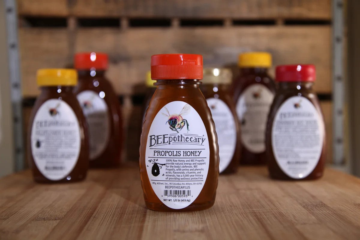 BEEpothecary propolis honey in 0.5 lb clear squeeze jar with red cap. Honey is deep amber. Additional honey containers are blurred out in the background.