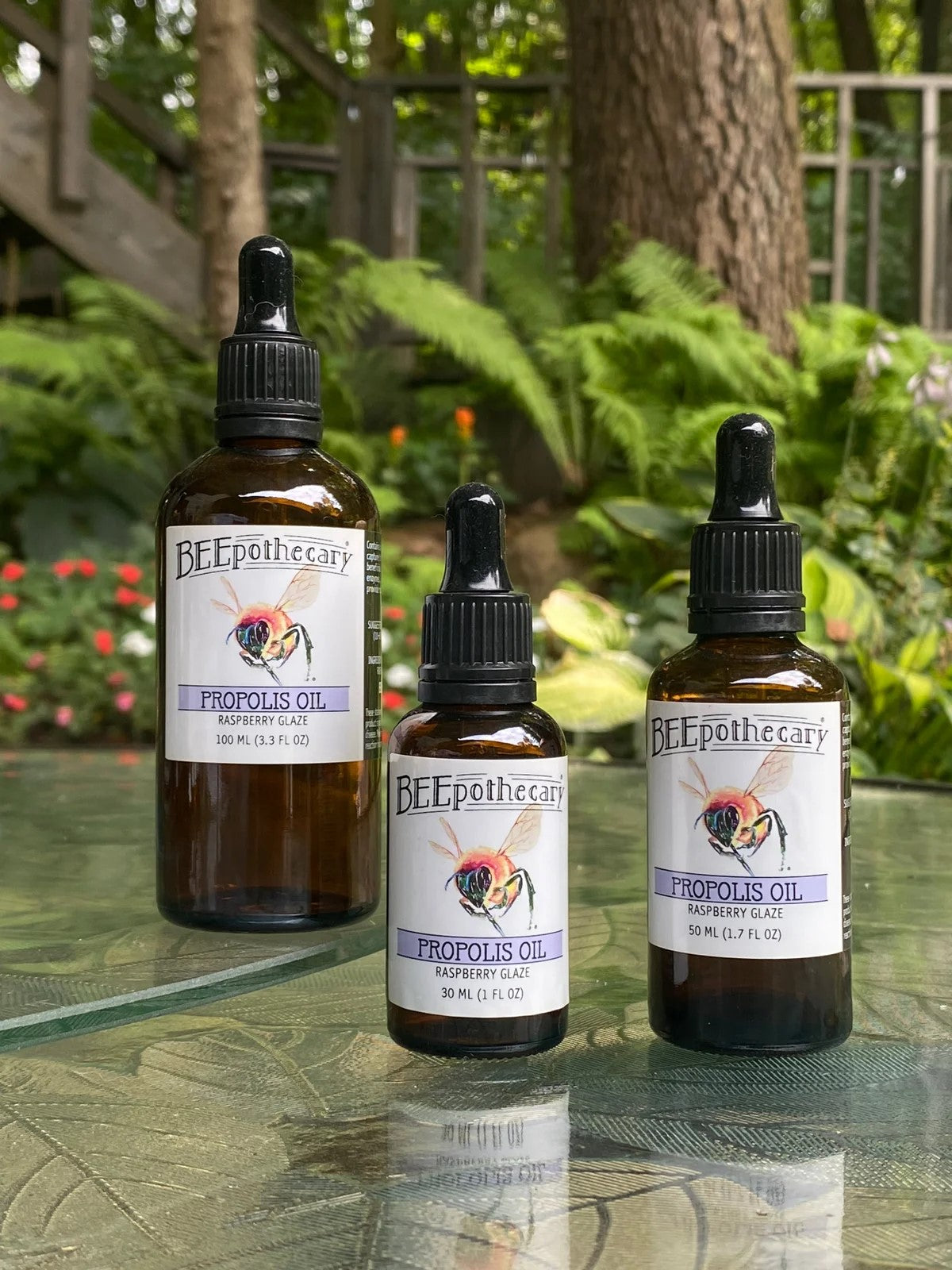 BEEpothecary Propolis Oil Raspberry Glaze in 30 ml, 50 ml, 100 ml brown bottles with white label and dropper displayed outdoors.