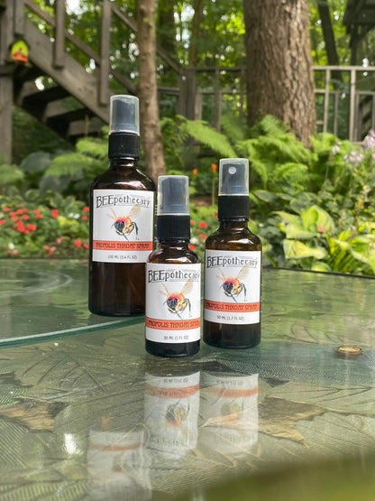 BEEpothecary Propolis Throat Spray in 30 ml, 50 ml, 100 ml brown spray bottles with white label displayed on table outdoors.