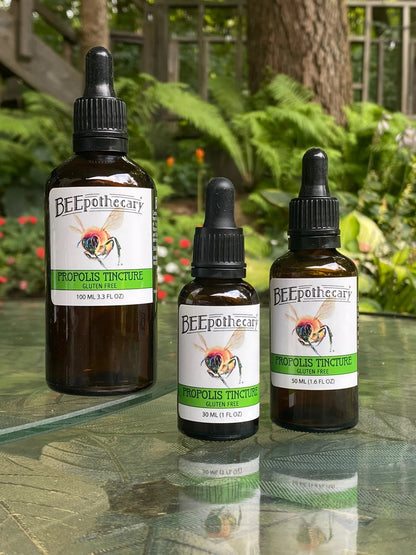 BEEpothecary Propolis Tincture Gluten Free in 30 ml, 50 ml, 100 ml brown bottles with white label and dropper displayed on table outdoors.